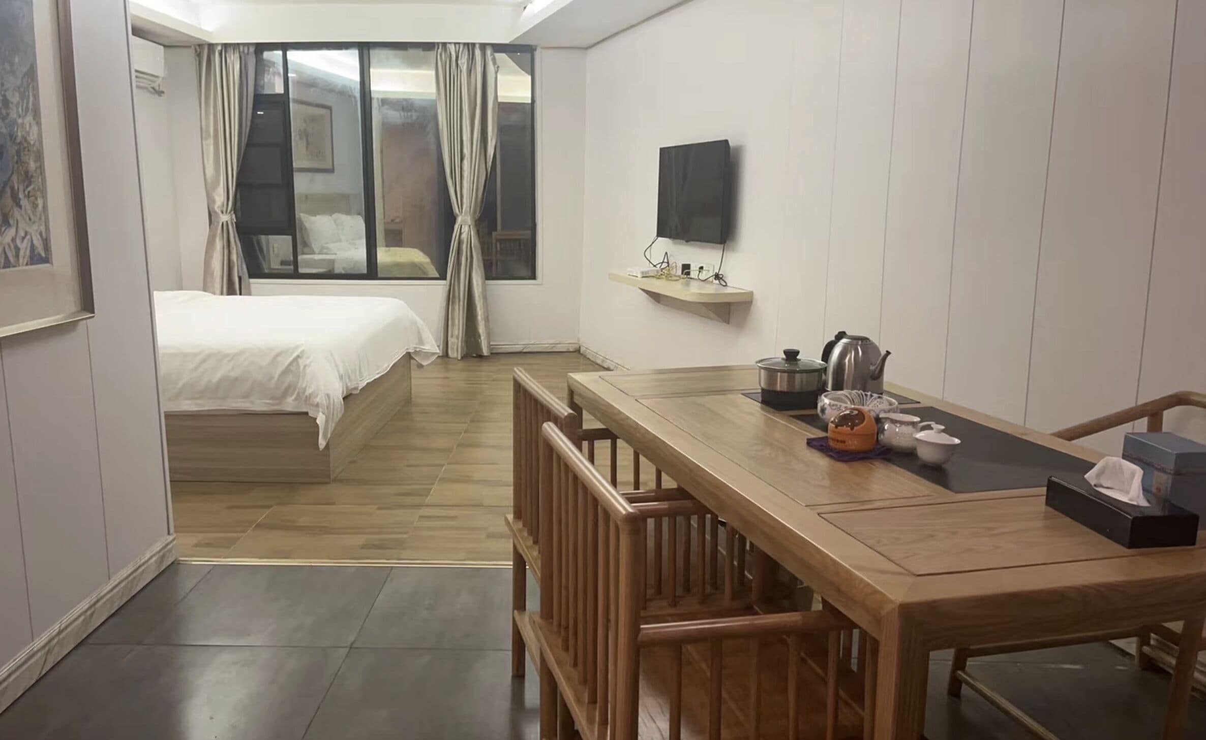 A hotel room with a wooden dining table, a bed, a TV mounted on the wall, and a small shelf with kitchen items.