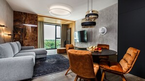Una sala de estar moderna con un sofá seccional gris, una mesa de comedor redonda con sillas, un televisor de pantalla plana montado en la pared y un amplio ventanal con vista a follaje.