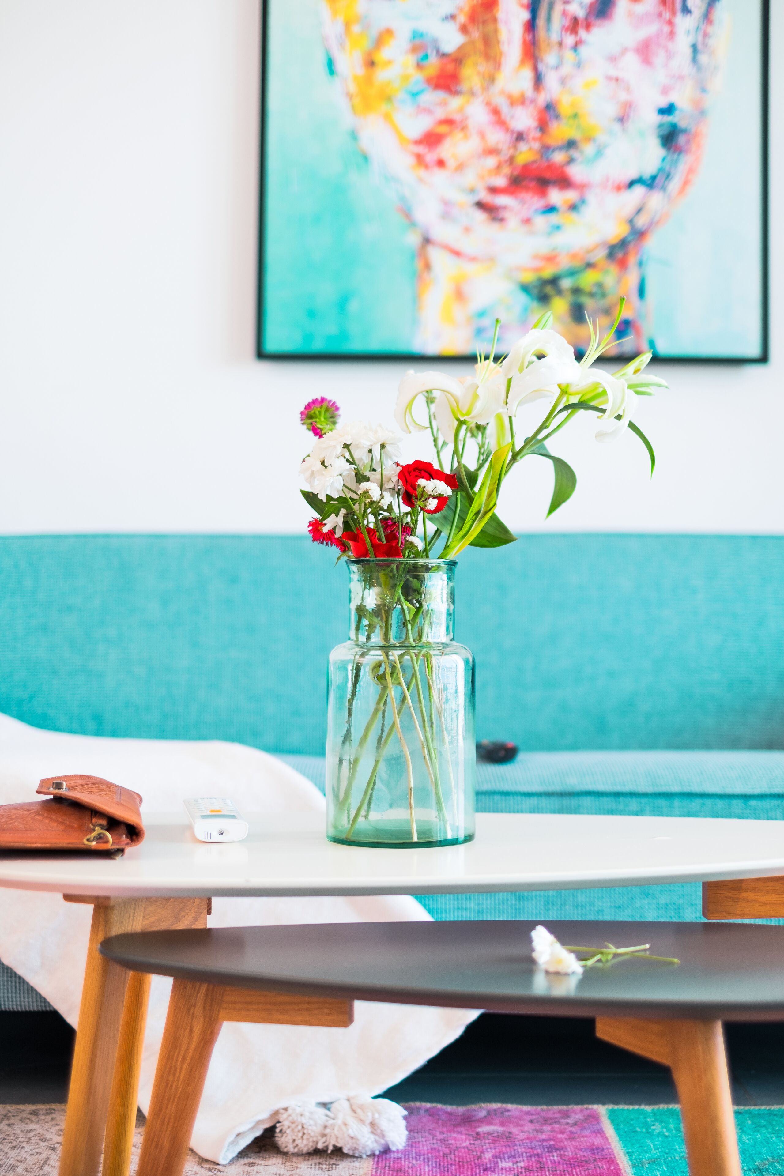 Un vase en verre transparent, orné de fleurs, est posé sur une table blanche dans une pièce aux murs de couleur turquoise, avec une peinture abstraite et colorée en arrière-plan.