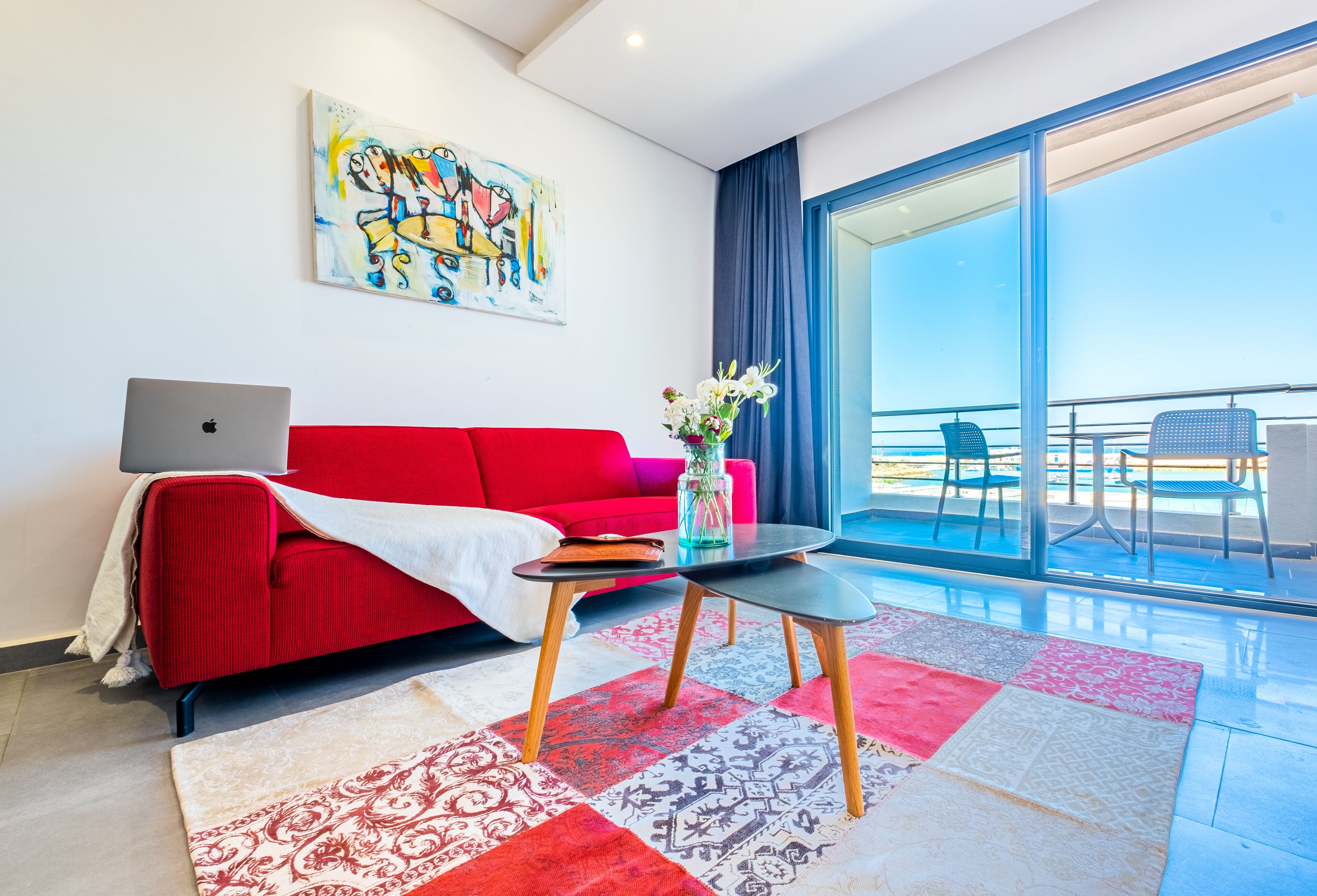 Una sala de estar moderna con un sofá rojo, un ordenador portátil sobre una mesita auxiliar y un gran ventanal con vistas a un balcón amueblado.