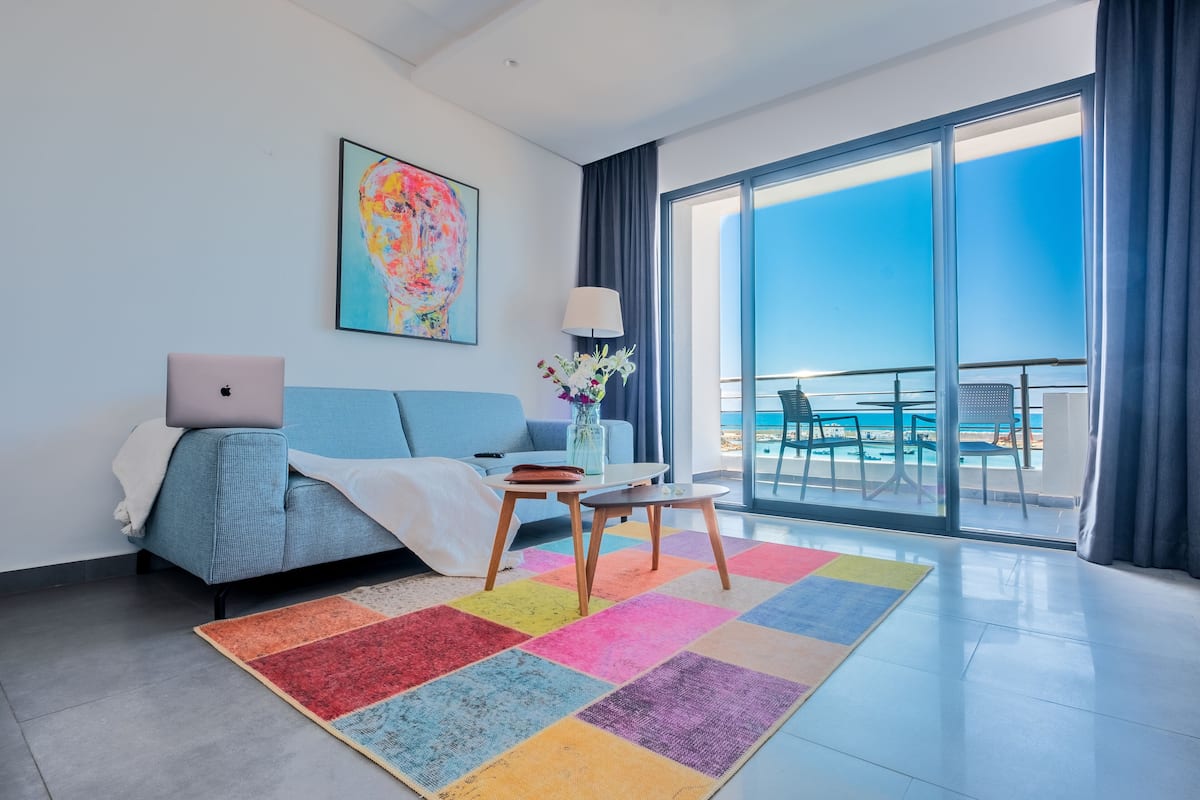 Una habitación de hotel moderna con un balcón grande, un ordenador portátil sobre la cama, una alfombra colorida y un cuadro en la pared.