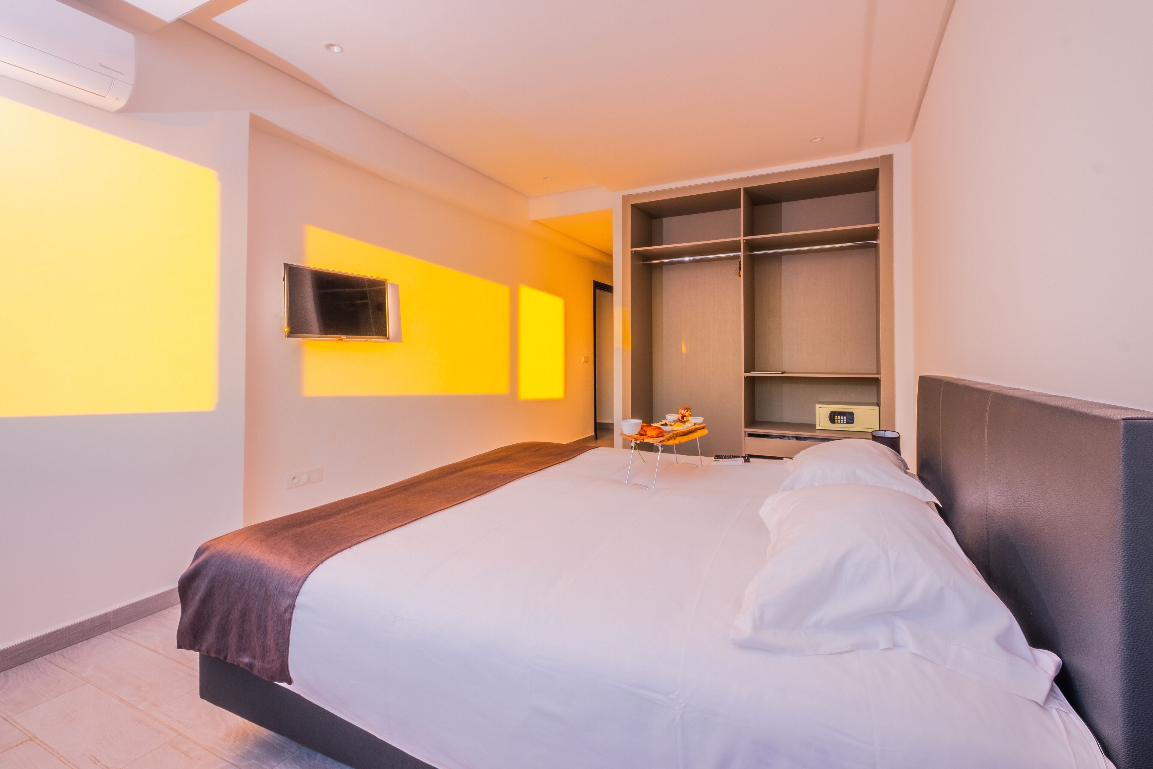 A modern hotel room with a large bed, a bedside table with a lamp, a TV mounted on the wall, and a wardrobe with shelves.