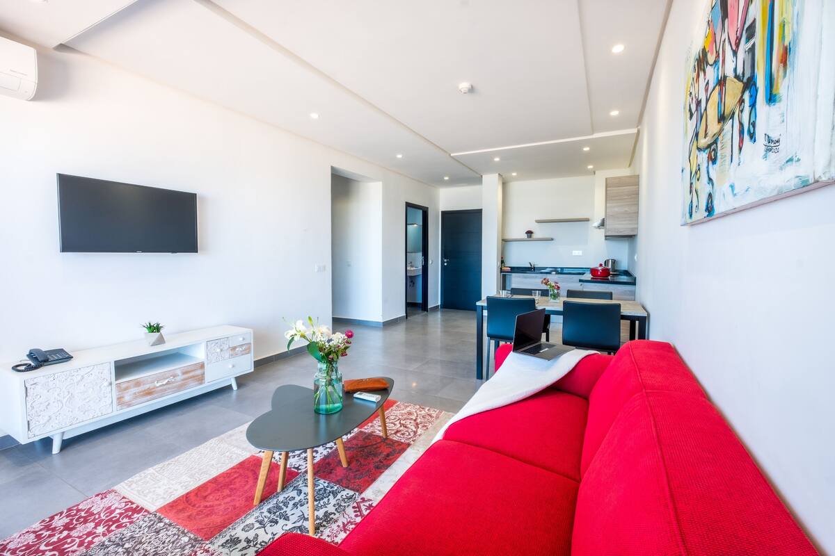 Una sala de estar moderna con un sofá rojo, un televisor de pantalla plana, un mueble bajo blanco y un comedor al fondo.