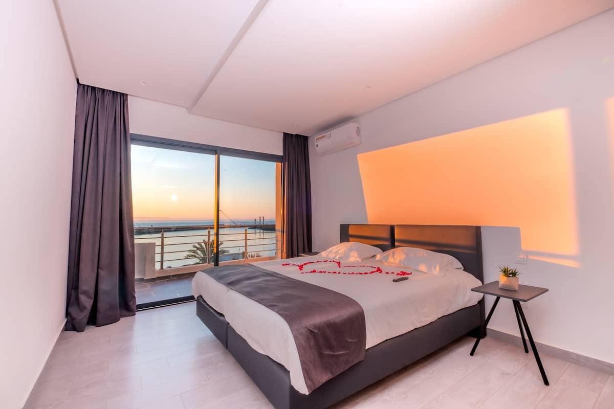 Una habitación de hotel moderna con una cama grande, una mesita de noche pequeña y vistas a la puesta de sol sobre el agua.