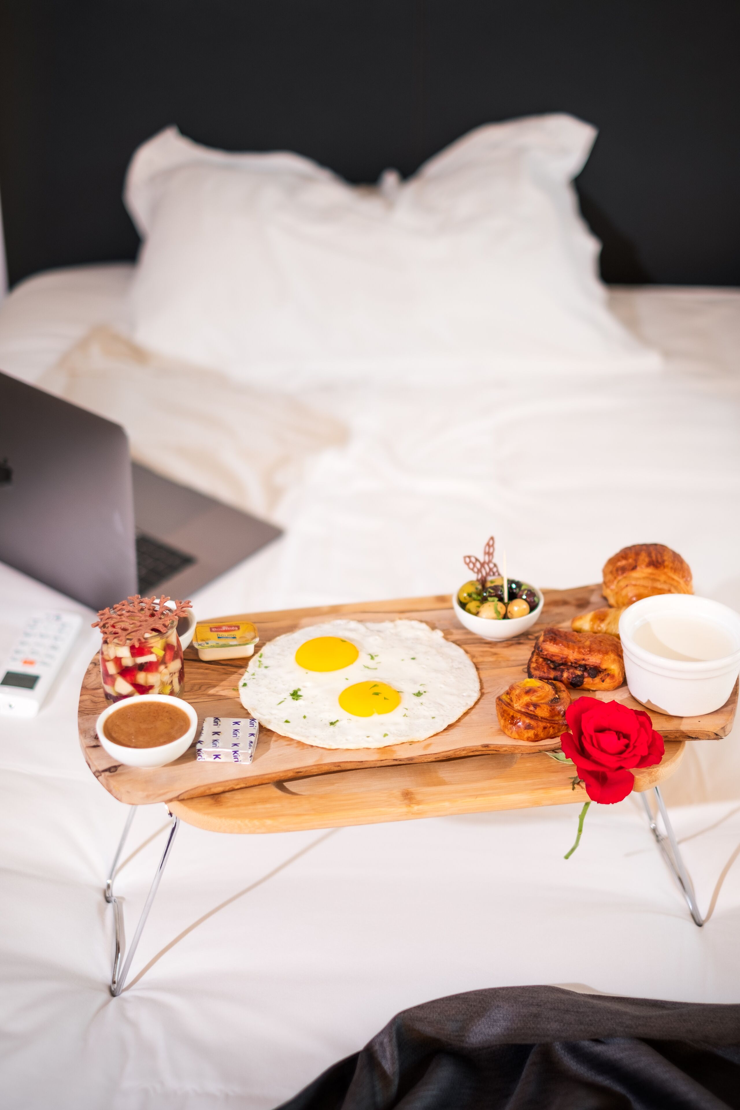 A breakfast tray with eggs, pastries, and a coffee cup on a bed with a laptop and a rose.
