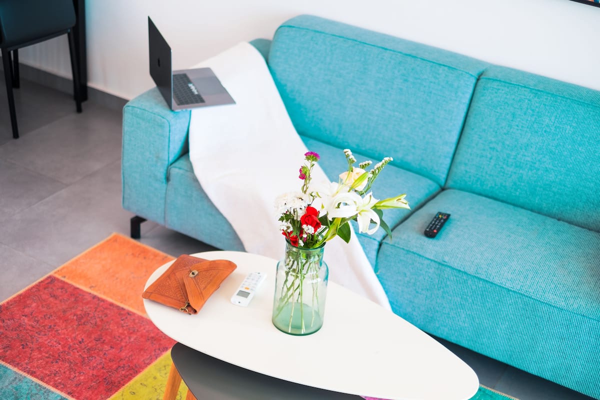 Un sofá azul aguamarina con un ordenador portátil y un mando a distancia encima, una almohada blanca, una alfombra colorida, un florero de cristal con flores y un bolso marrón sobre una mesa blanca.