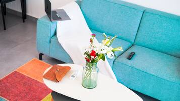Um sofá verde-turquesa com um laptop e um controle remoto sobre ele, uma almofada branca, um tapete colorido, um vaso de vidro com flores e uma bolsa marrom em uma mesa branca.