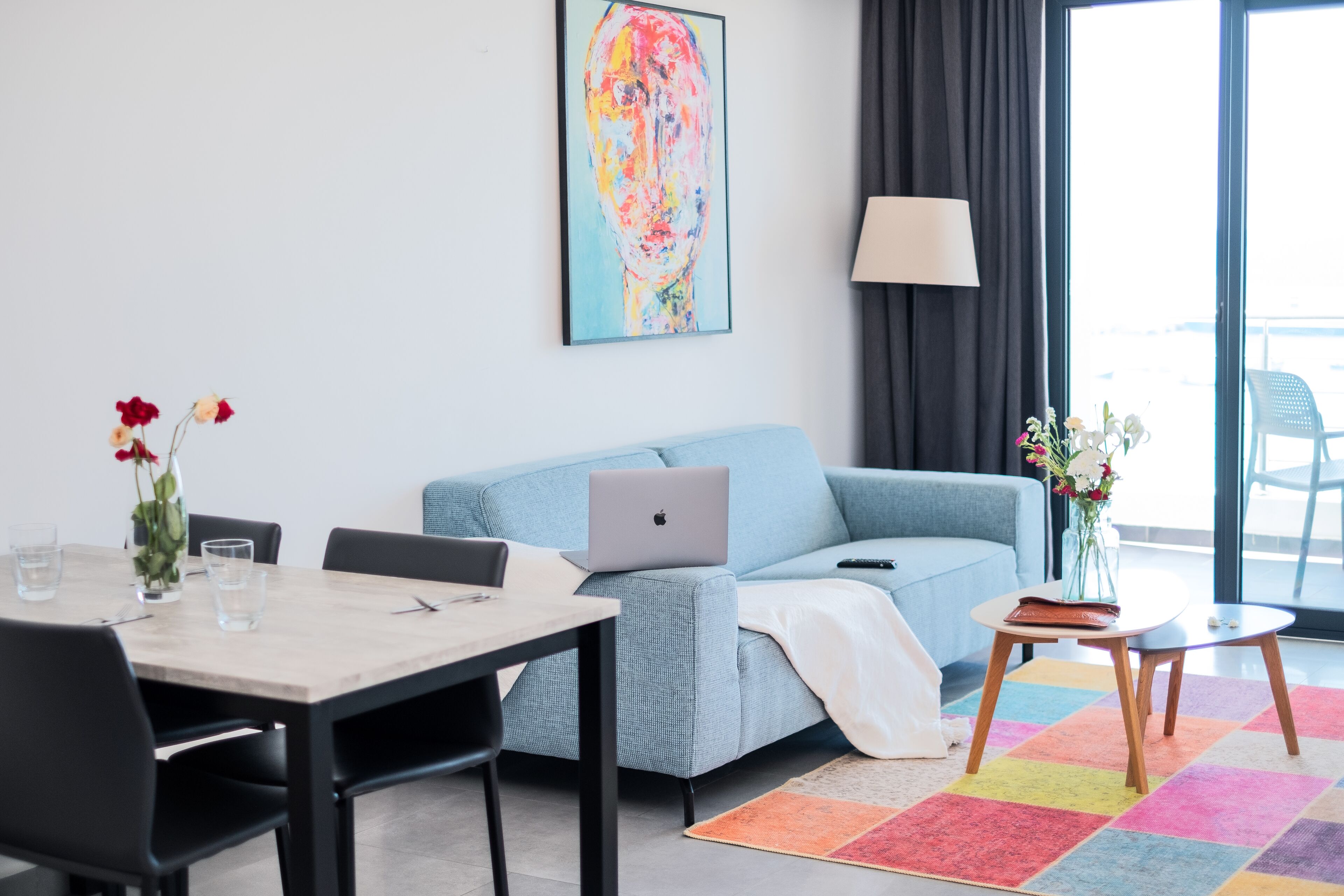 A modern living room with a light blue sofa, a laptop on the sofa, a dining table with chairs, a colorful rug, and a painting on the wall.
