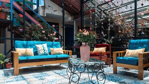 Un patio con suelo azul estampado, un sofá de madera con cojines decorados, una silla de madera y una mesa de vidrio con un jarrón de flores.