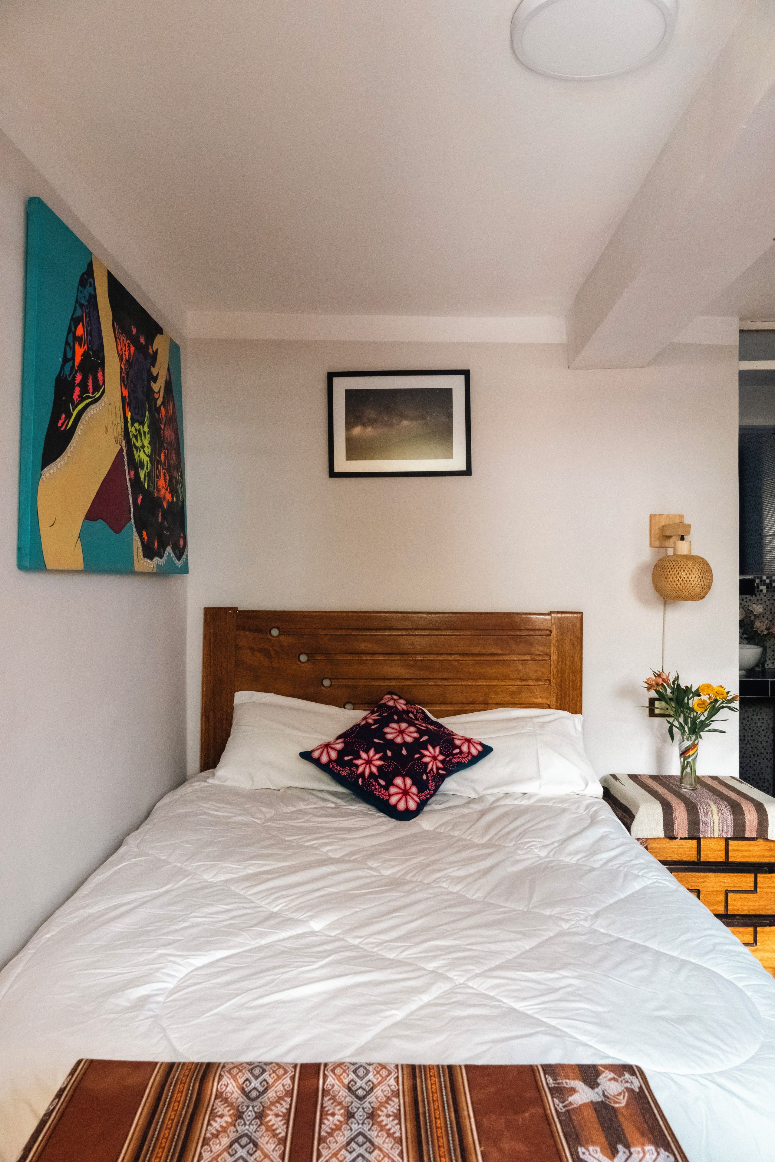 Quarto com cabeceira de madeira, colcha branca, uma almofada decorativa, quadro na parede e uma pequena mesa com um vaso de flores.