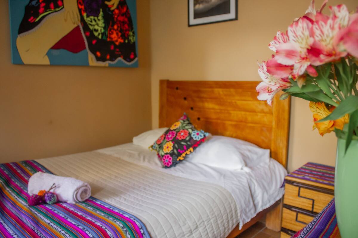 Una cama con edredón blanco, manta colorida y almohada estampada. Se observa un cabecero de madera, una mesita de noche con mantel decorado y un florero con flores.
