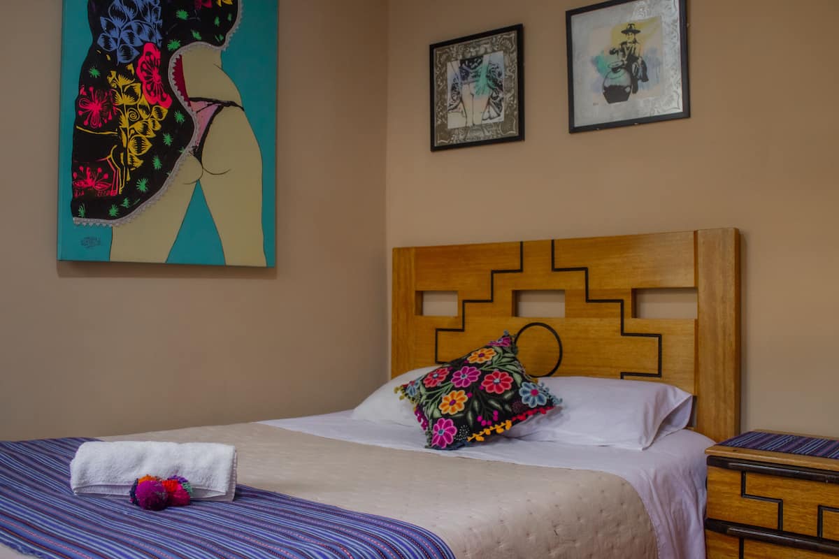 Habitación de hotel con una cama, cabecera de madera, colcha a rayas, almohada blanca, almohada con estampado floral y una toalla blanca. Dos cuadros en la pared.