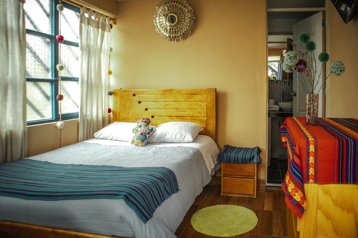 Un dormitorio con cabecera de madera, cama con ropa blanca, mesita de noche, una pequeña mesa con mantel colorido y una ventana con cortinas.