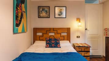 Un dormitorio con cabecera de madera, una cama con manta azul, dos cuadros enmarcados en la pared y una mesita de noche con lámpara.