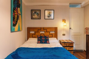 Un dormitorio con cabecera de madera, una cama con manta azul, dos cuadros enmarcados en la pared y una mesita de noche con lámpara.