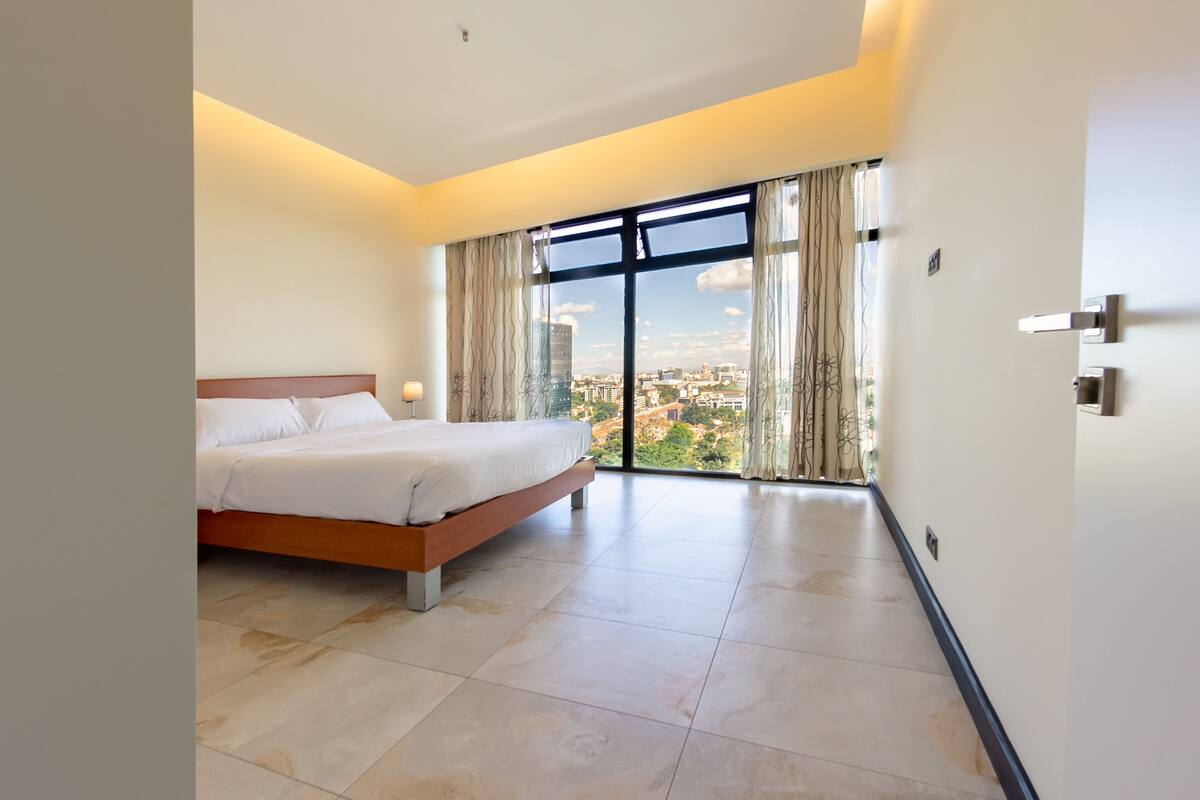 Una habitación de hotel moderna con una cama grande, vistas de la ciudad a través de ventanales del suelo al techo, y un diseño limpio y minimalista.