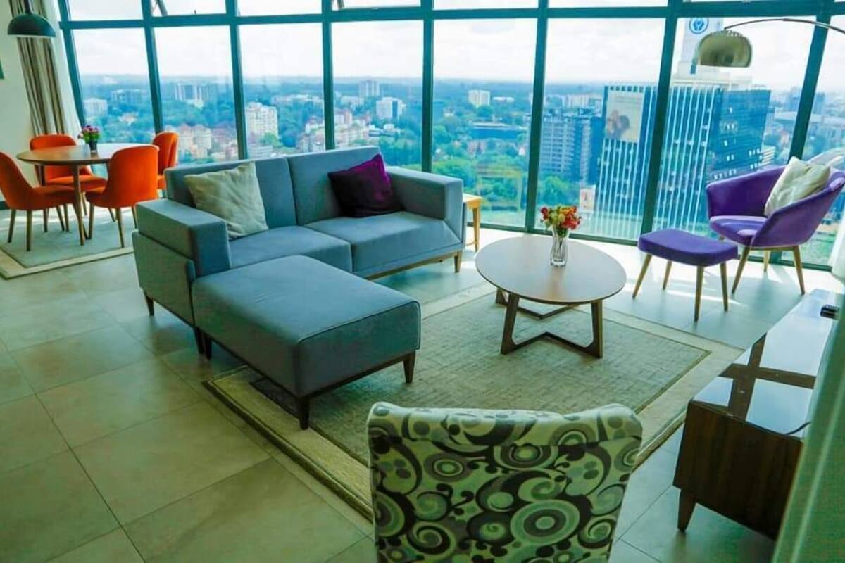 Una sala moderna con un sofá seccional gris, un sillón estampado y una mesa de centro redonda. Hay una zona de comedor con sillas naranjas y una mesita. La habitación tiene ventanas grandes con vistas a la ciudad.