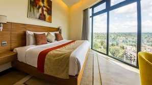 Een moderne hotelkamer met een groot bed, een houten hoofdbord, een schilderij aan de muur en uitzicht op de stad door grote ramen.
