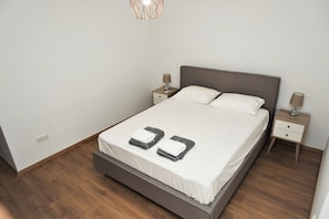 A neatly made bed with white linens and two folded towels, a wooden floor, and two bedside tables with lamps. - Koules Sea View City Apartment (Heraklion)