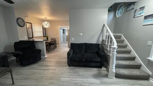 Amplia sala de estar con un sofá de color oscuro, una mesa de comedor con sillas y una escalera con balaustres blancos.