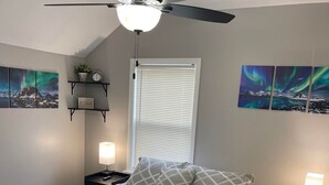 Un dormitorio con cama, ventilador de techo, lámparas de noche, una ventana con persianas y decoración en la pared.
