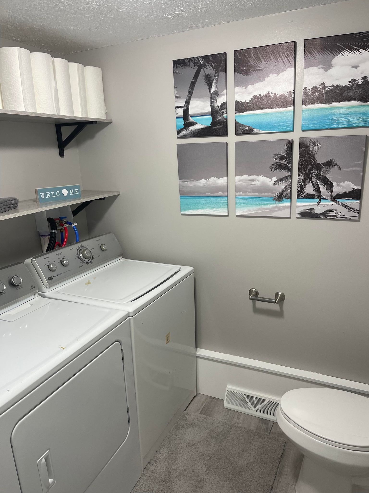 A laundry room with a washing machine, dryer, and a shelf with paper towels. There are four framed pictures on the wall depicting tropical beach scenes.