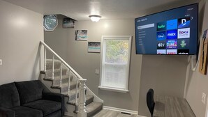 Una sala de estar con un sofá gris, una mesa de madera, una silla de oficina negra, una escalera y un televisor en la pared que muestra la interfaz de una plataforma de streaming.