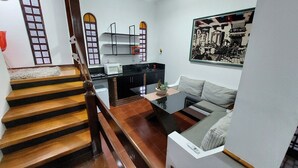 A modern kitchen with a black countertop, a built-in oven, and a microwave. There is a staircase with wooden steps leading to a landing. A dining area with a glass table and a sofa is visible. - Amplo estudio proximo ao BOS em Sorocaba (Sorocaba)