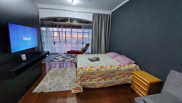 Quarto com cama, televisão, poltrona e vista para o exterior através de uma janela.