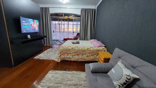 A modern bedroom with a bed, a grey sofa, a television, and a view of the city through large windows. - Amplo estudio proximo ao BOS em Sorocaba (Sorocaba)
