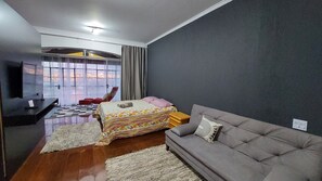 Quarto moderno com cama, sofá, televisão e uma janela grande com cortinas.