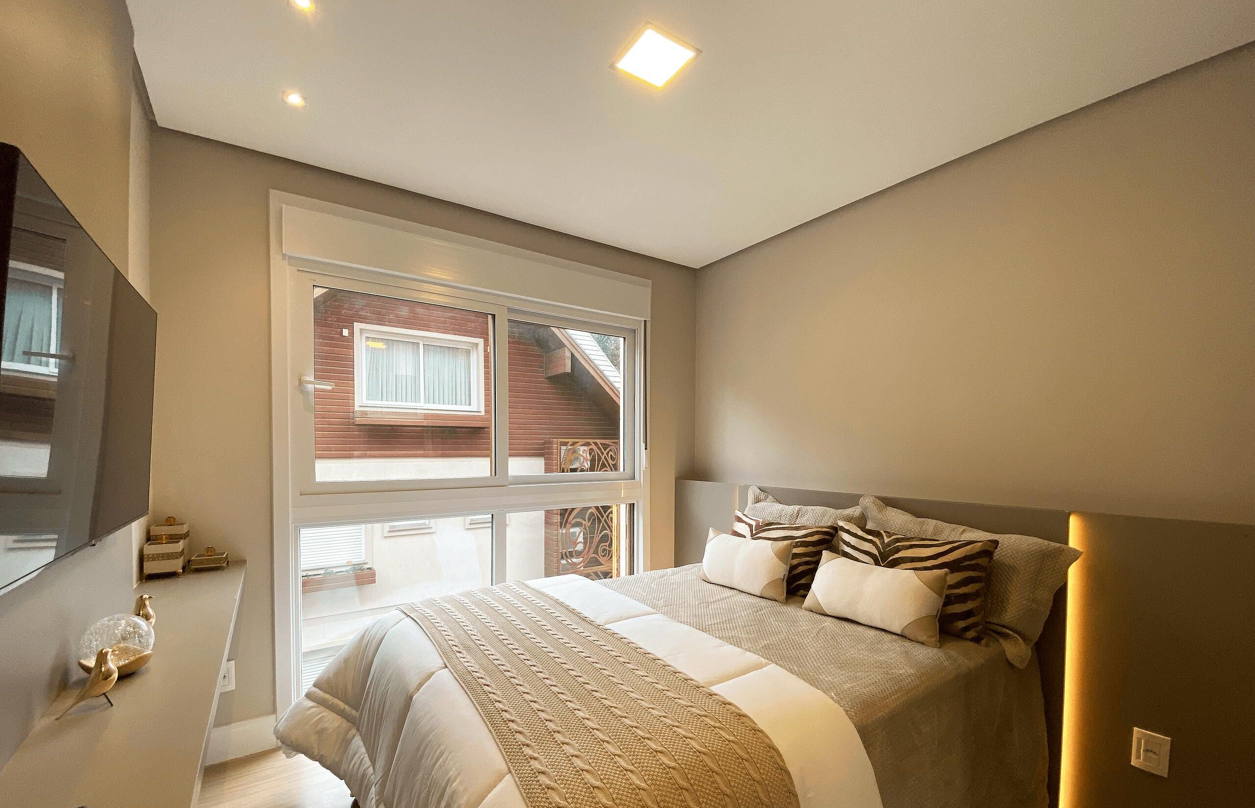 Un dormitorio moderno con una cama grande, un televisor montado en la pared y un ventanal con vistas a un edificio de ladrillo.