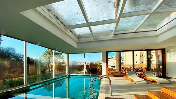 Una piscina interna moderna con pareti in vetro trasparente, un solarium in legno e poltrone relax.