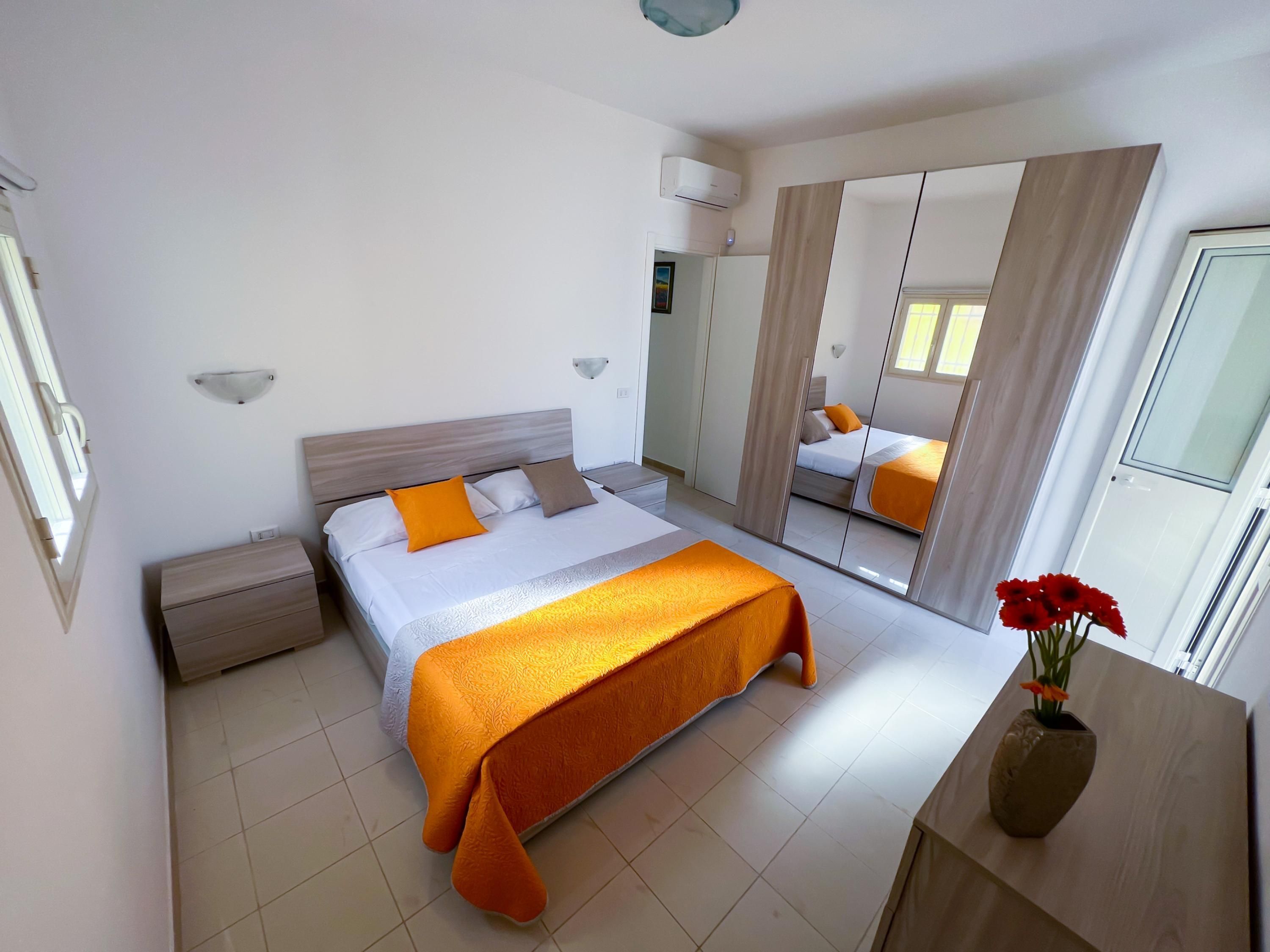 A modern hotel room with a large bed, a bedside table, a mirror, and a vase with flowers.