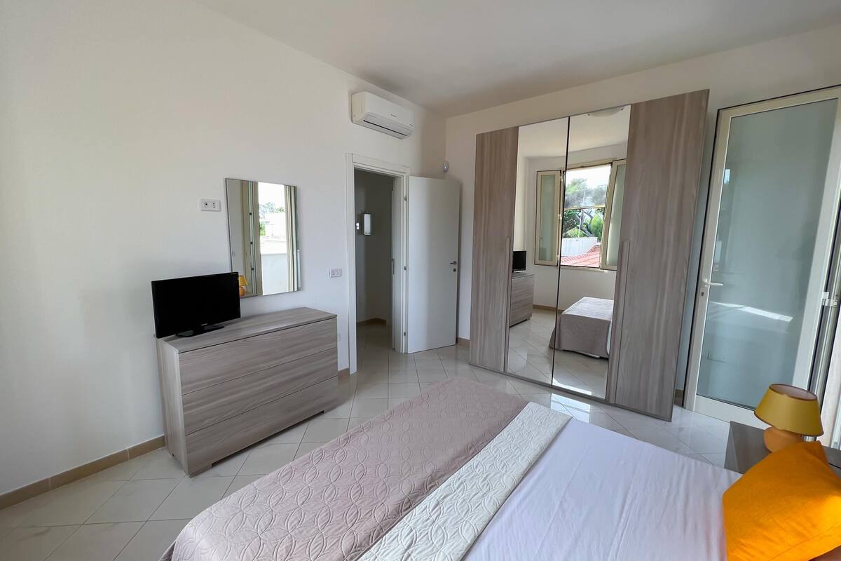Ein modernes Schlafzimmer mit einem Bett, einer Kommode, einem Fernseher und einer Glastür, die auf einen Balkon führt.