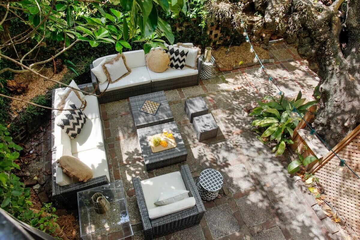 Un espace extérieur accueillant, aménagé avec un canapé, deux tables carrées et une table ronde, le tout entouré de verdure et d’un mur en pierre.