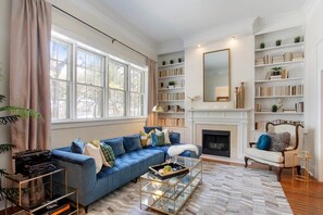 A living room with a blue sofa, a fireplace, bookshelves, and a large window.