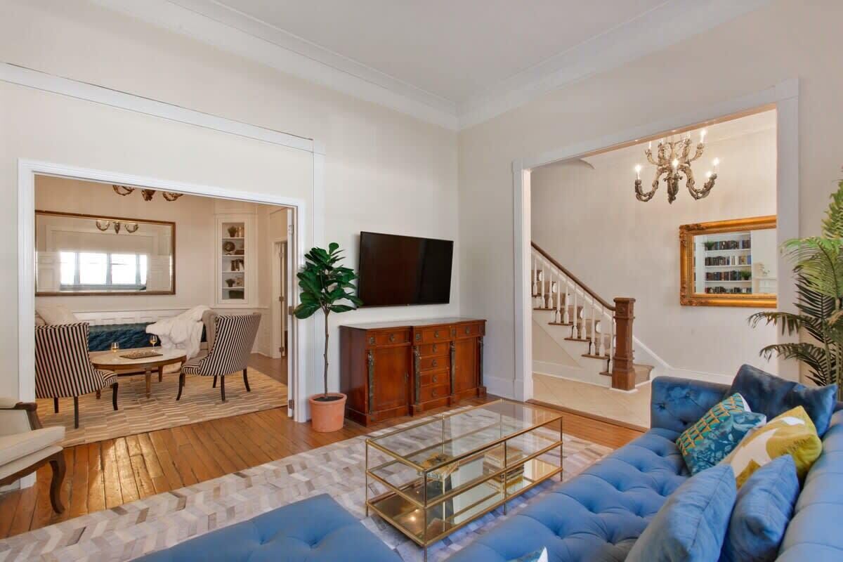 Ein großzügiges Wohnzimmer mit einem blauen Sofa, einem Glastisch und einem Fernseher an der Wand. Eine Treppe mit einem Kronleuchter und einem großen Spiegel darüber.