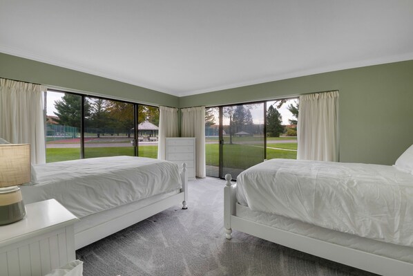 Un dormitorio con dos camas, un aparador y amplias ventanas que ofrecen vistas a un campo deportivo.