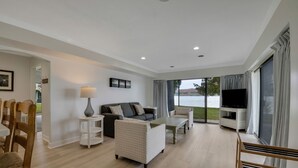A modern living room with a sofa, coffee table, and TV, large windows with curtains, and a view of the outdoors.