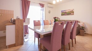 Een eetgedeelte met een glazen tafel, zes roze stoelen, een wit kastje en een schilderij aan de muur.