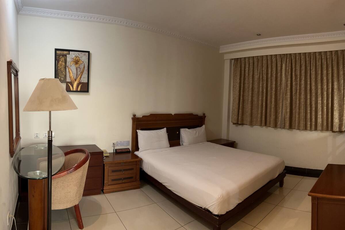 Una habitación de hotel con cama, escritorio con lámpara, una silla y un cuadro en la pared.