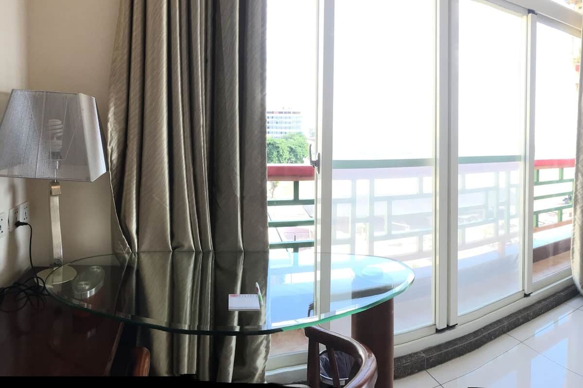 Habitación de hotel con una mesa de vidrio, televisión, refrigerador y balcón con cortinas.