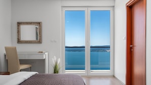 Een slaapkamer met een bed, een bureau, een stoel, een groot raam met uitzicht op zee en een houten deur.