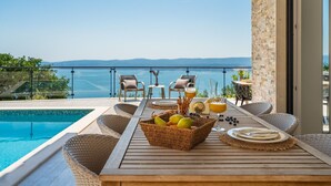Een houten tafel met een fruitmand, een glas sinaasappelsap en een waterfles op een balkon met uitzicht op een zwembad en een schilderachtig zeegezicht.