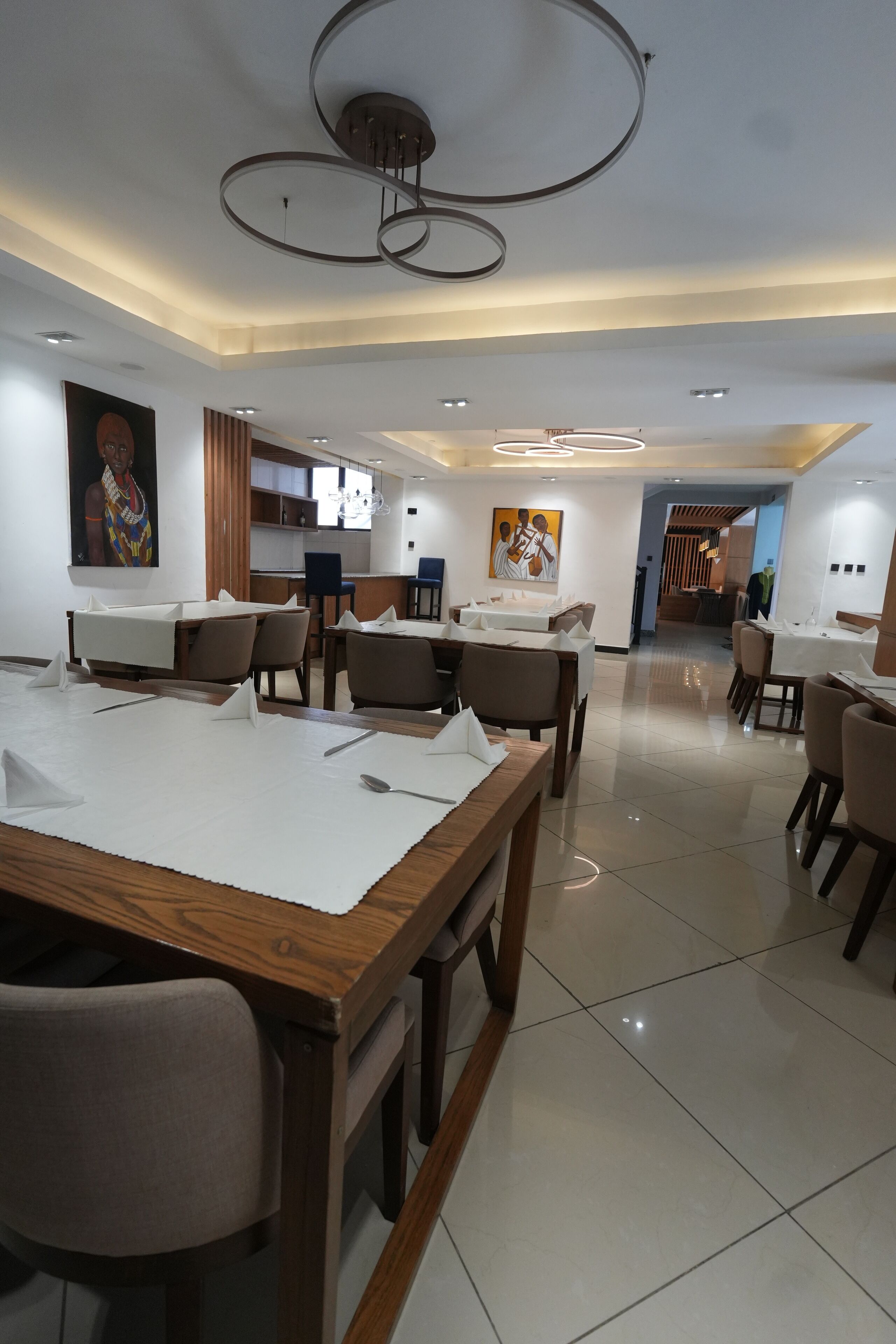 A modern dining area with multiple tables and chairs, a kitchen in the background, and a ceiling with circular light fixtures.