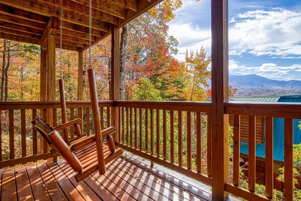 Una terraza de madera con una mecedora, con vistas a un bosque de follaje otoñal y montañas distantes.