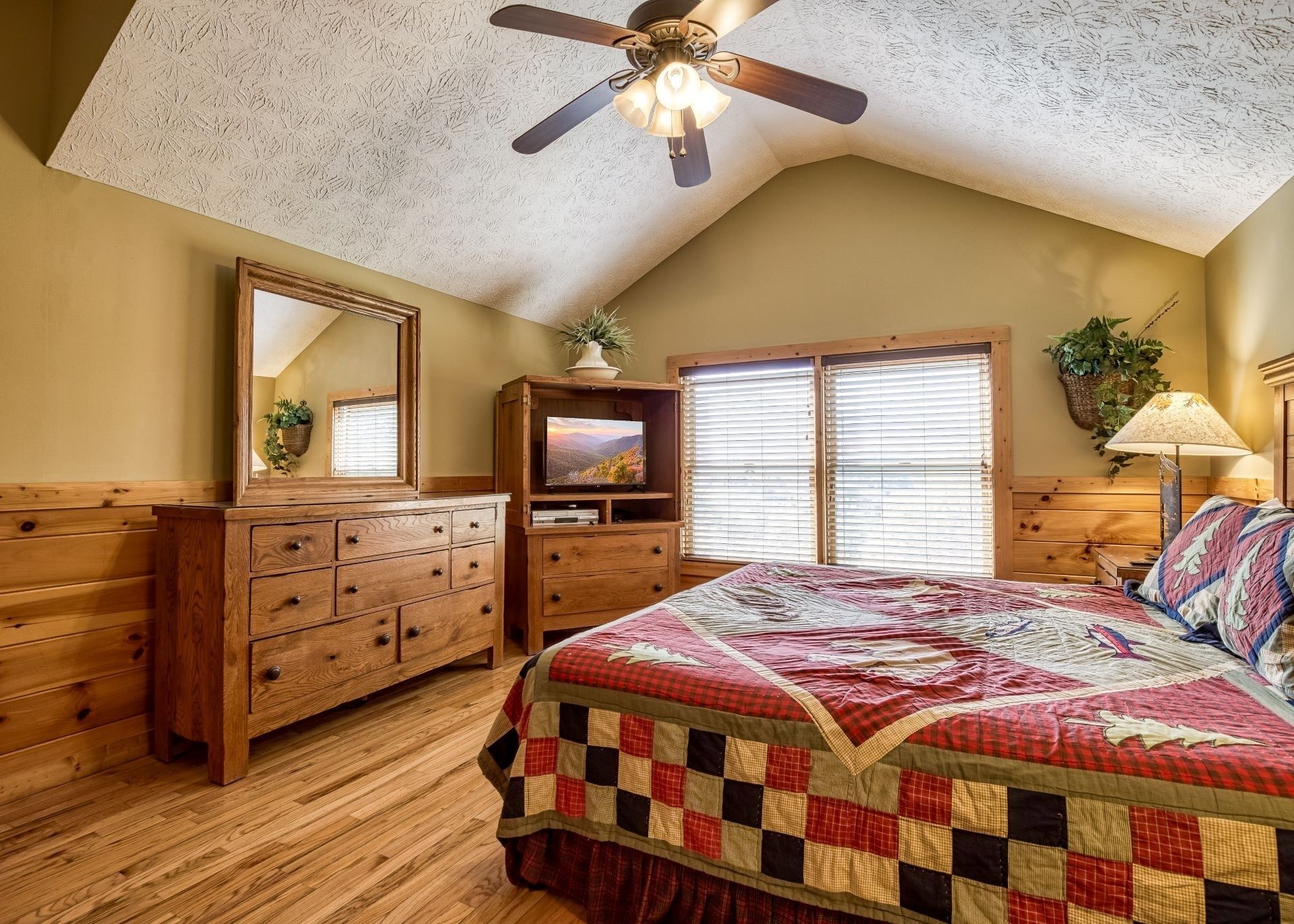 A bedroom with a wooden dresser, a bed with a quilt, a ceiling fan, and a television on a wooden cabinet.