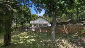 A two-story house with a large front porch, surrounded by a stone wall and trees. - Tavi Haus: Lakefront~Kayaks~SUP~Sauna~Pool Table (Empire)