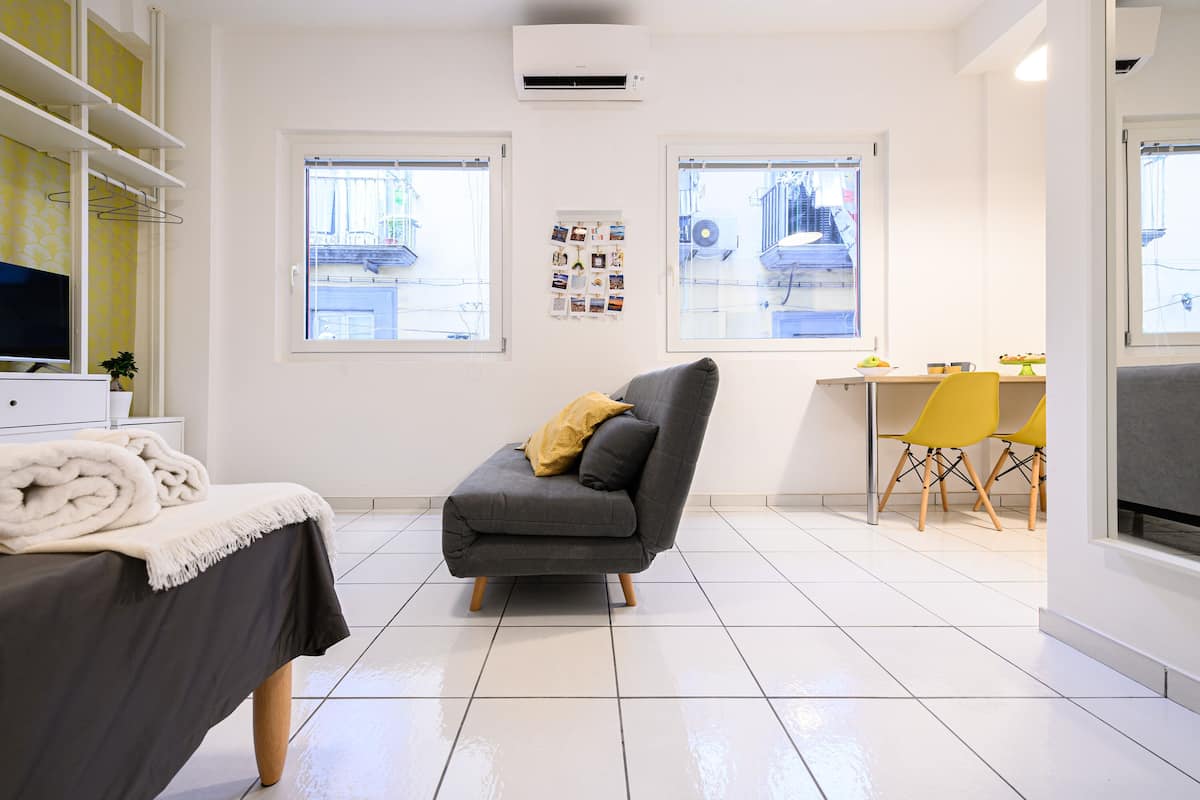 Une pièce moderne avec un canapé gris, une table à manger avec des chaises jaunes et un téléviseur posé sur un meuble blanc.